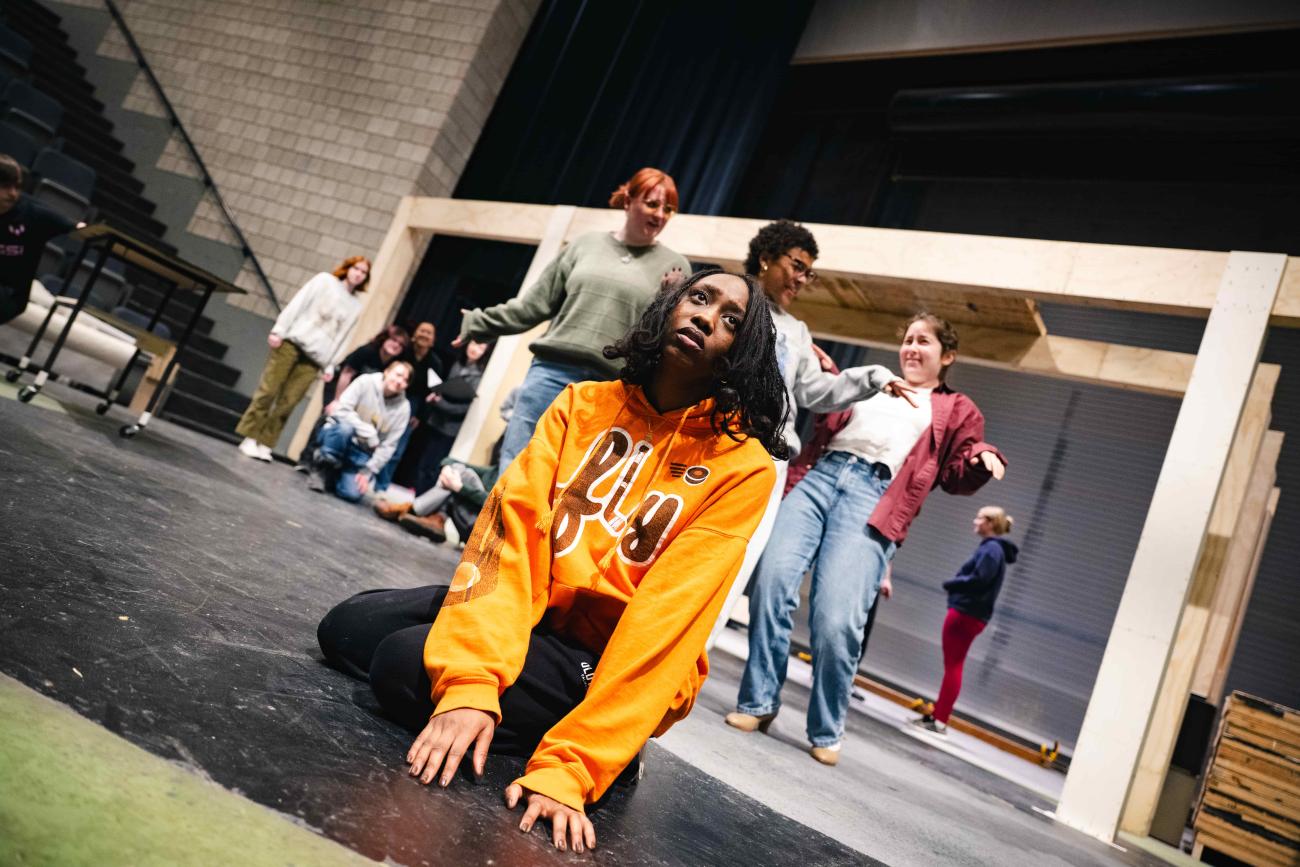 Actress sitting on the floor looking concerned wearing a yellow fly sweatshirt while others are dancing behind her.