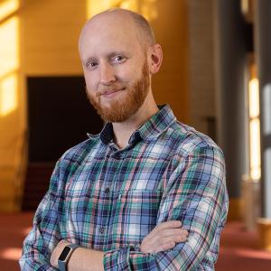 Will Lowry wearing a plaid shirt, smiling at the camera with his arms crossed in Zoellner Arts Center's lobby.
