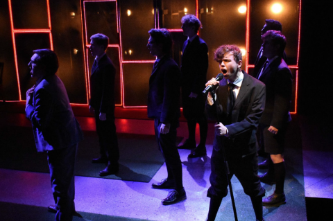 A person dressed in a black suit singing into a microphone surrounded by other singers.