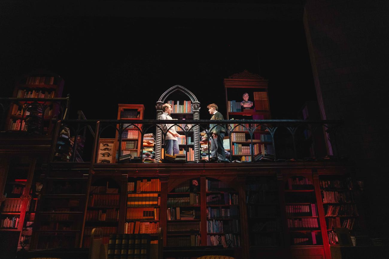 Actors talking on a the second floor balcony of a bookshelf.