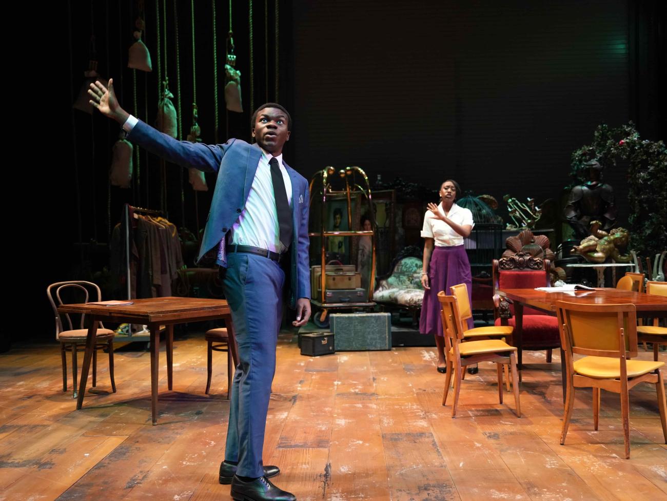 Actor dressed in a suit talking with his right arm raised, while an actress talks to him from behind.