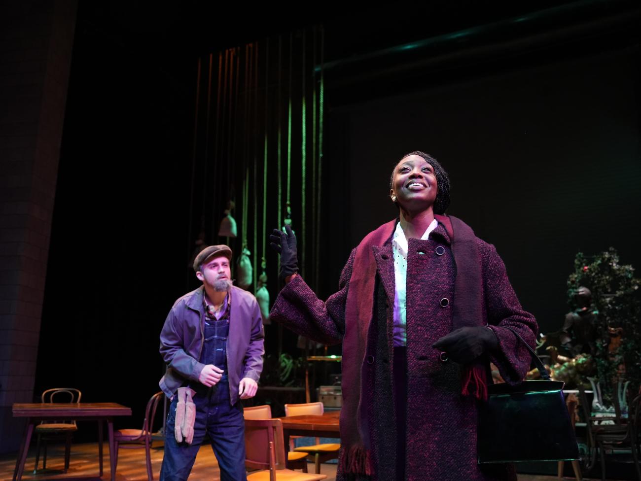 Actress wearing a coat looking up the ceiling with an actor standing behind her wearing a hat.