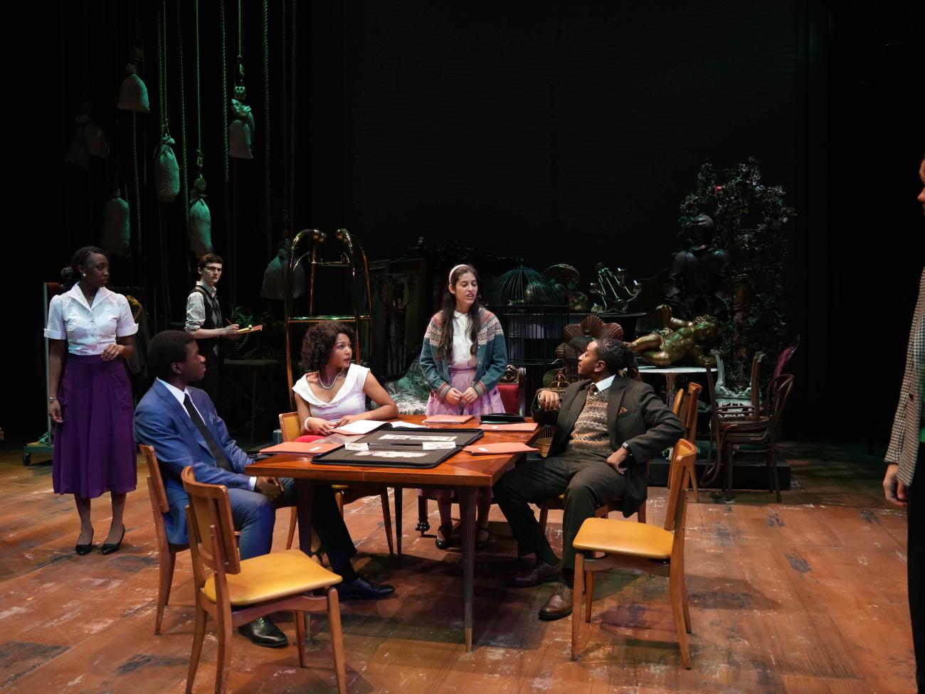 Actors sitting around a table looking at an actress while she standing and talking.