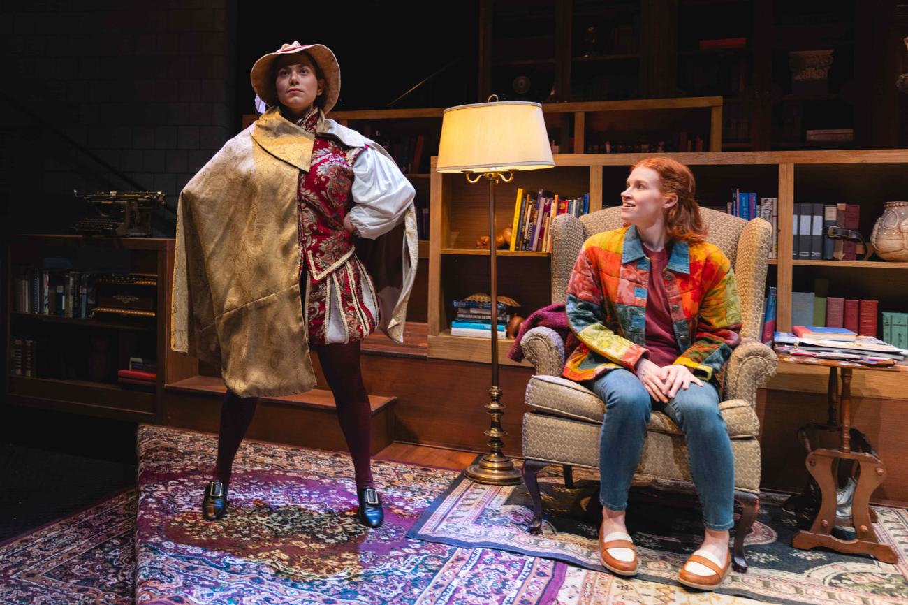 Actress dressed in a costume with his hand on her hip standing next to another actress sitting on a chair.