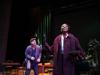 Actress wearing a coat looking up the ceiling with an actor standing behind her wearing a hat.