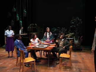 Actors sitting around a table looking at an actress while she standing and talking.