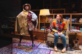 Actress dressed in a costume with his hand on her hip standing next to another actress sitting on a chair.