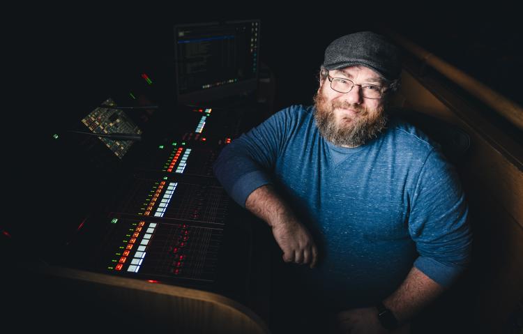 Phillip Ingle in a blue shirt and hat sitting in front of audio controls