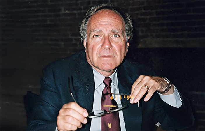 Jeff Milet wearing a suit and tie holding glasses looking at the camera.