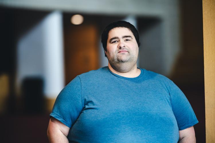 Justin Burns wearing a blue t-shirt standing for his portrait in a hallway.