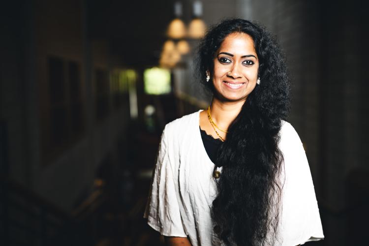 Swapna Suresh wearing a white shirt and black undershirt smiling for portrait in a dark hallway with a light shining on her.
