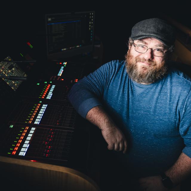 Phillip Ingle in a blue shirt and hat sitting in front of audio controls