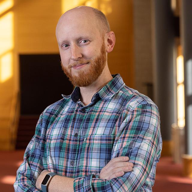 Will Lowry wearing a plaid shirt, smiling at the camera with his arms crossed in Zoellner Arts Center's lobby.