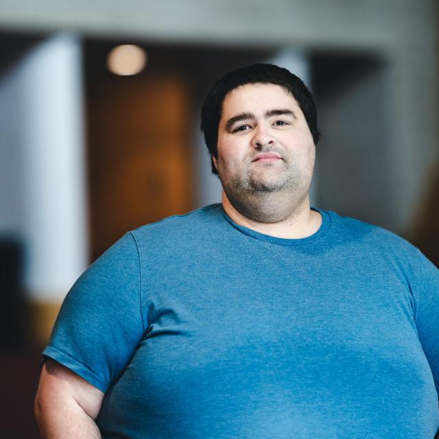 Justin Burns wearing a blue t-shirt standing for his portrait in a hallway.