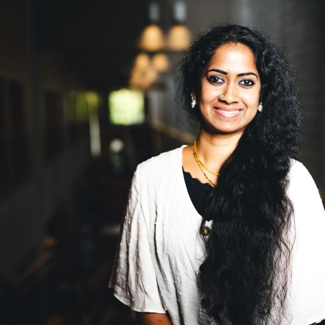 Swapna Suresh wearing a white shirt and black undershirt smiling for portrait in a dark hallway with a light shining on her.