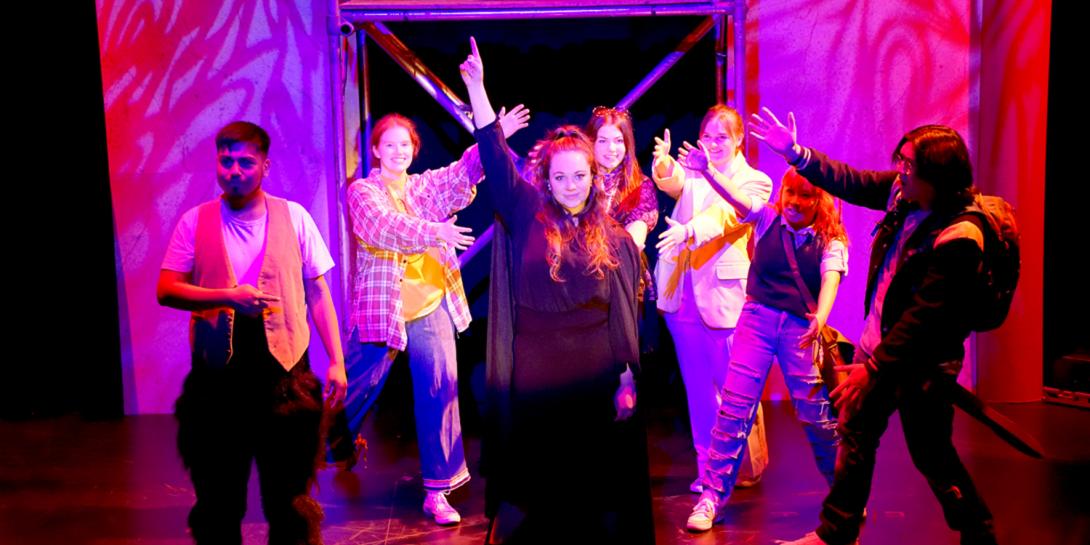 A group of actors in a neon pink room with an actress pointing up to the sky.