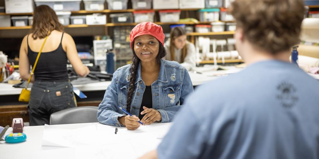 A female student holding a pencil to draw on paper and smiling at a male student.