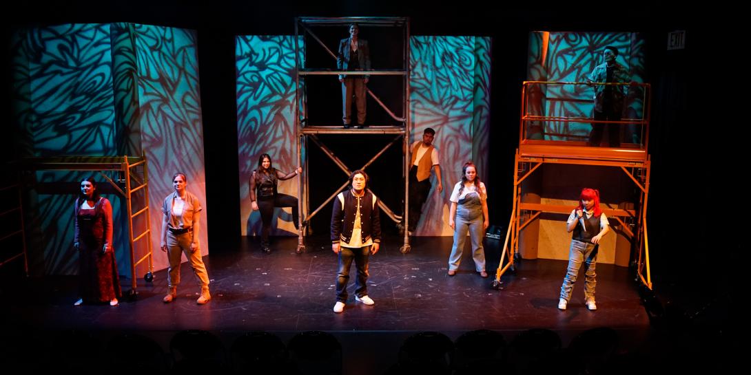 9 actors standing in front of scaffolding looking at the audience.