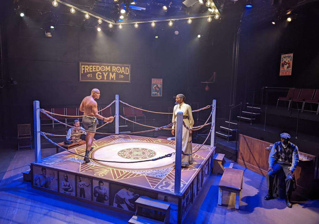 A male in shorts and boxing gloves with a woman in a yellow dress in a boxing ring facing each other inside a gym called Freedom Road Gym..