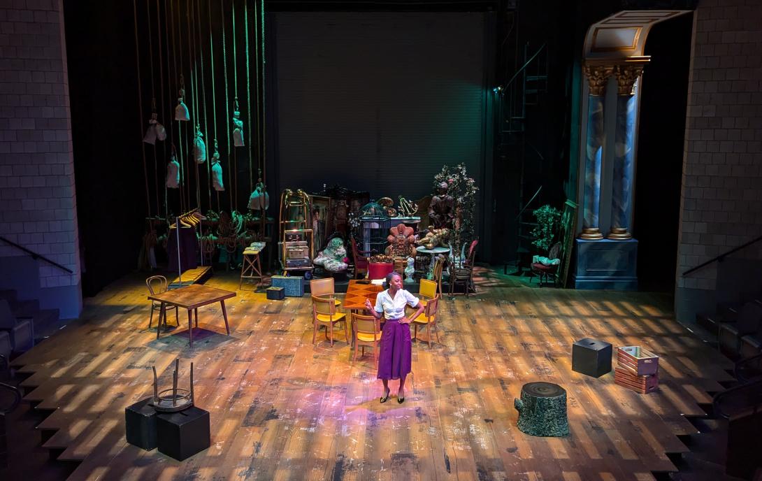 A female wearing a pink skirt and white blouse standing with her hand on her hip with furniture and artifacts in the background.